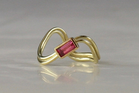 A gold ring with a twisted, openwork band and a rectangular pink gemstone set in the center, displayed on a light gray reflective surface.