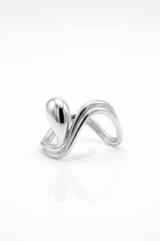 A polished silver ring with a sculptural, flowing wave design and a rounded teardrop-shaped element on top, displayed on a clean white reflective surface.
