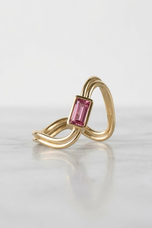 A gold ring with a twisted, layered band and a rectangular pink gemstone, displayed on a light gray reflective surface.