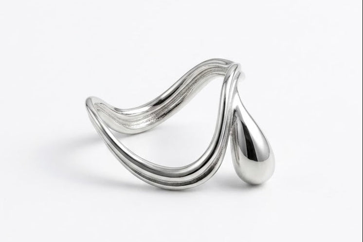 A polished silver-toned metal ring with a sculptural, flowing wave-like design and a teardrop-shaped end, photographed on a clean white background.