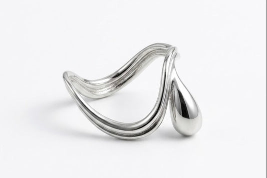 A polished silver-toned metal ring with a sculptural, flowing wave-like design and a teardrop-shaped end, photographed on a clean white background.