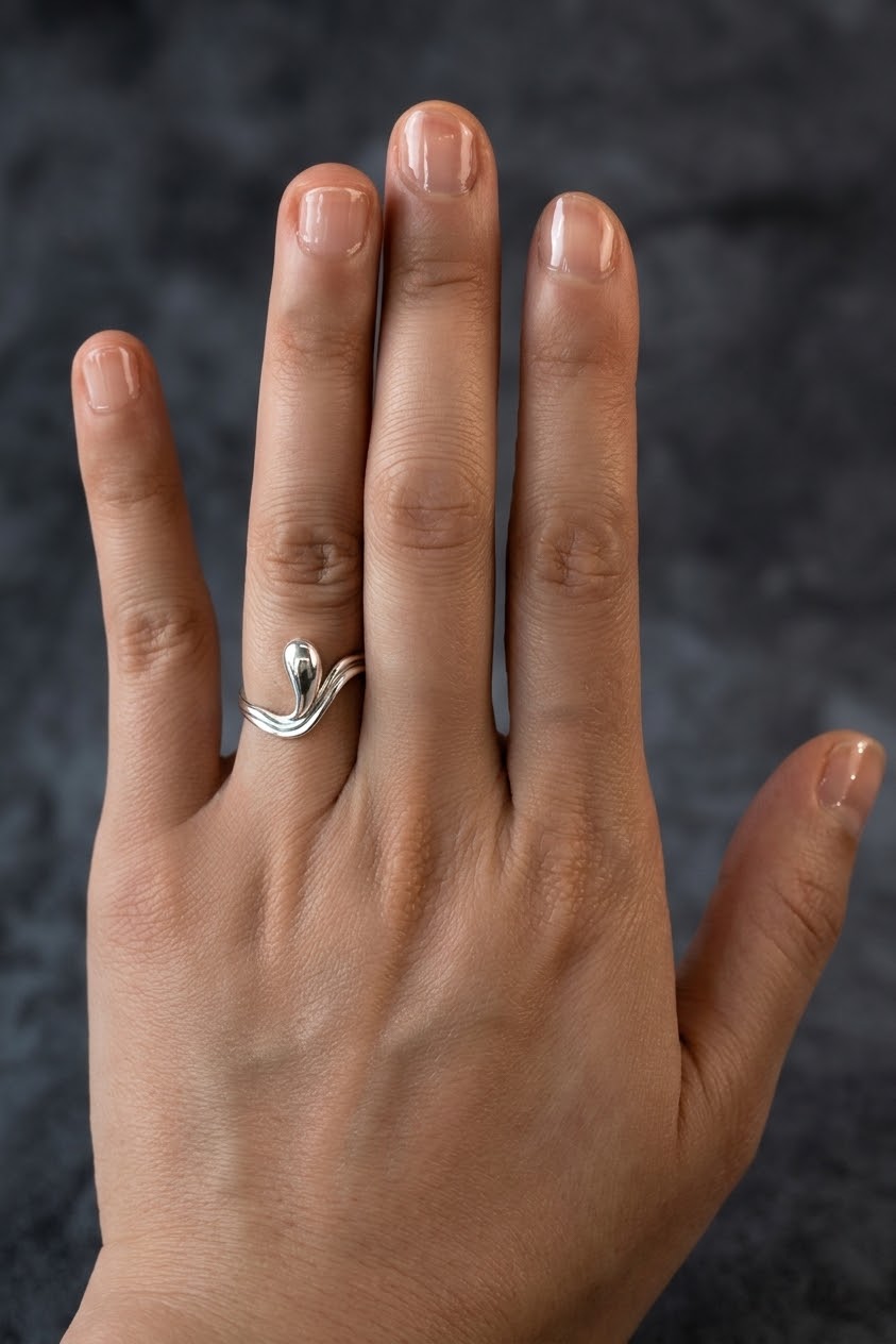 A close-up of a hand wearing a polished silver ring with a stylized, flowing wave or leaf-like design on the ring finger, set against a dark textured background.